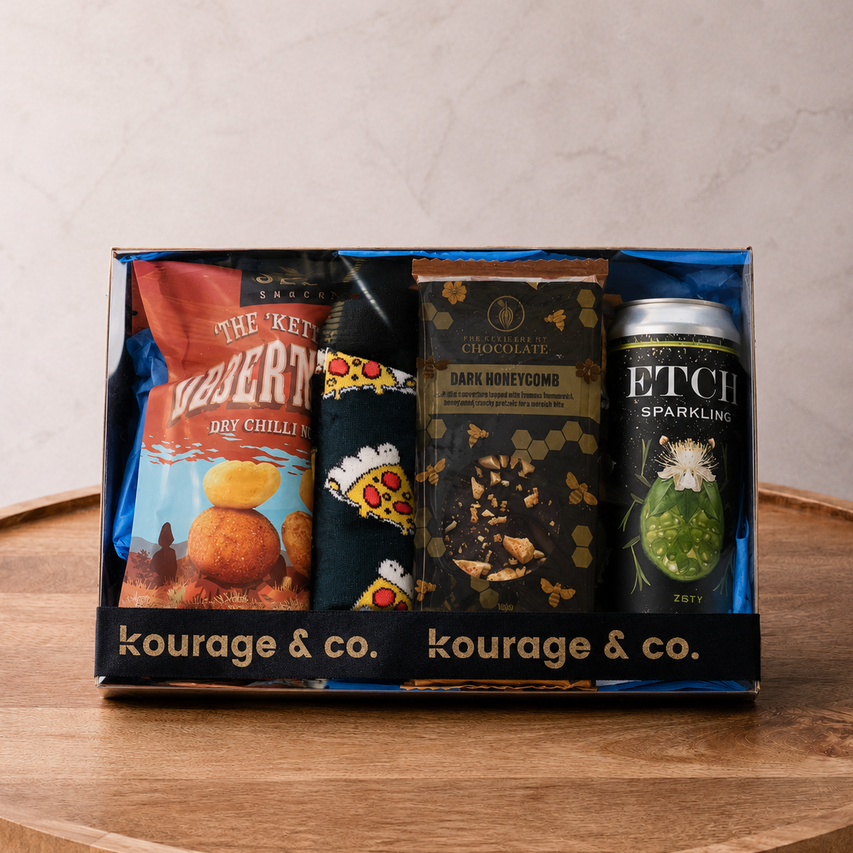 Gift box from kourage & co containing snacks and a can on a wooden surface.